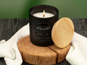 Candle in a black container with wooden lid on a wooden block against a green background